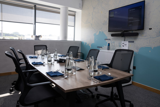 A conference room with a table set for a meeting, featuring bottles of water, glasses, and napkins. A large wall map and a screen are visible in the background.