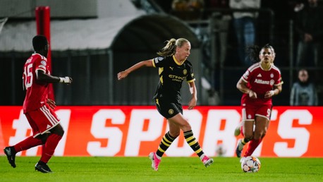 A football match with players in action. A player in a black and yellow kit controls the ball near opponents wearing red kits.