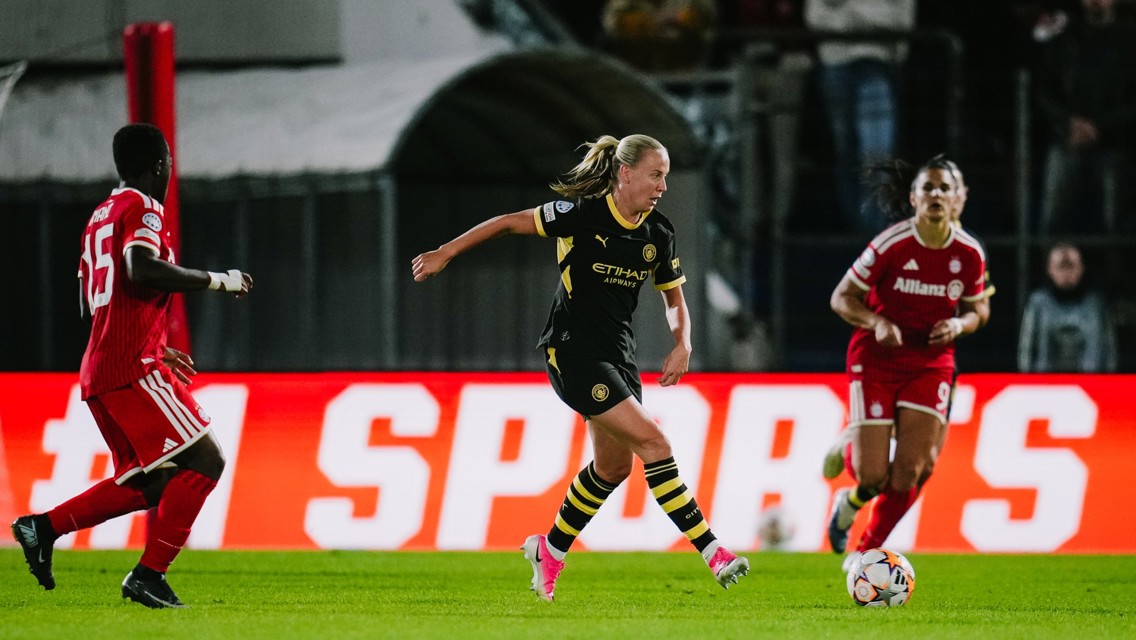 A football match with players in action. A player in a black and yellow kit controls the ball near opponents wearing red kits.