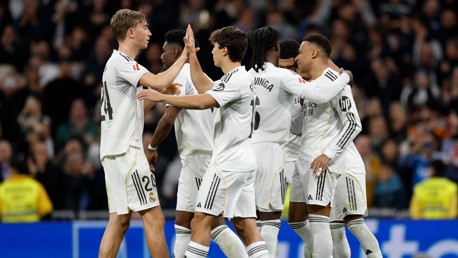 Real Madrid players celebrate a goal during a match, wearing white jerseys with logos and numbers visible. They are gathered together in a stadium, high-fiving and embracing each other.