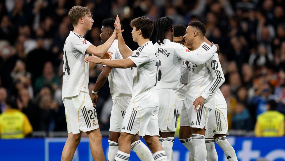 Real Madrid players celebrate a goal during a match, wearing white jerseys with logos and numbers visible. They are gathered together in a stadium, high-fiving and embracing each other.