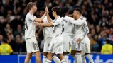 Real Madrid players celebrate a goal during a match, wearing white jerseys with logos and numbers visible. They are gathered together in a stadium, high-fiving and embracing each other.