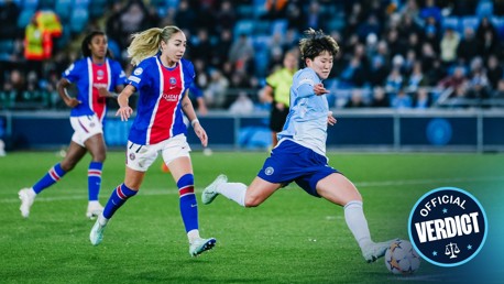 Soccer players in action during a match. One player in a light blue kit is kicking the ball. Two players in blue and red kits are behind and watching. Crowd in the background. 'Official Verdict' badge on the image.