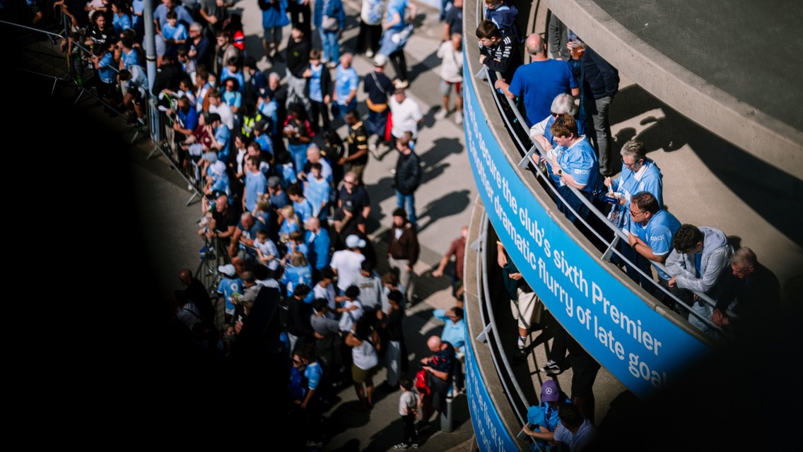 A large group of people wearing blue shirts gathered outside a stadium. Some are leaning against a railing with text about the club's achievements.