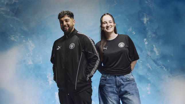 Two people in Puma Manchester City outfits, standing against a blue sky background; one wears a black jacket, the other a black shirt and blue jeans.