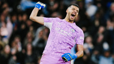 A football goalkeeper in a purple Manchester City kit celebrates with a clenched fist.