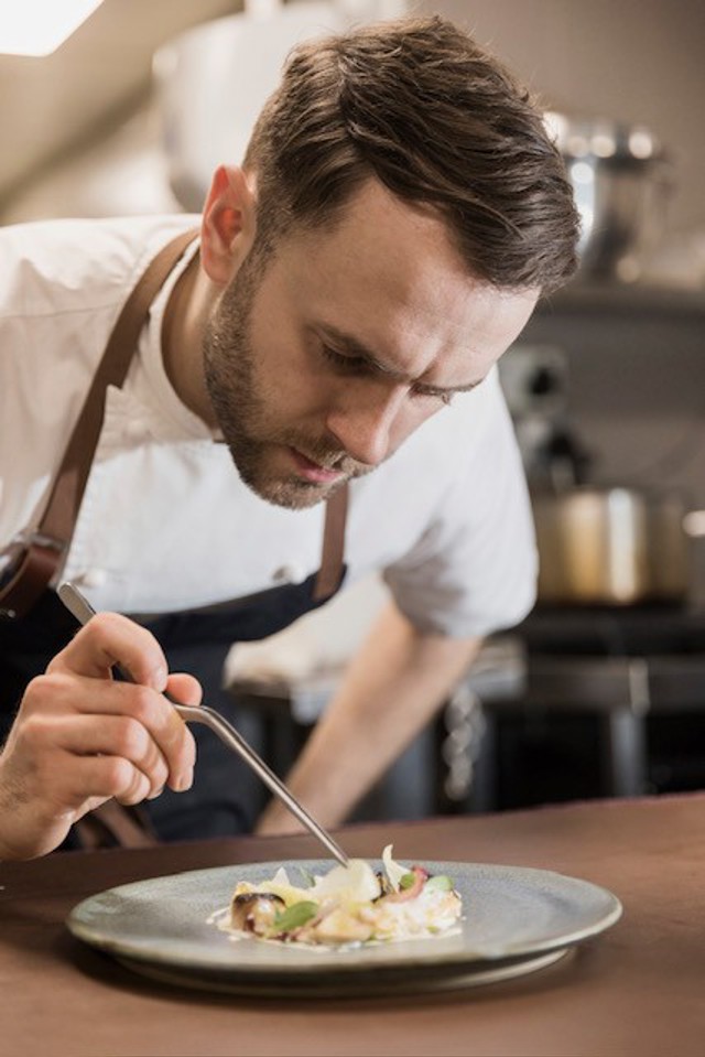 A chef is meticulously plating a dish in a professional kitchen, using a tool to arrange the food on a plate.
