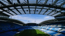 Interior view of a stadium with blue seats and a large roof structure, showing a clear sky above.