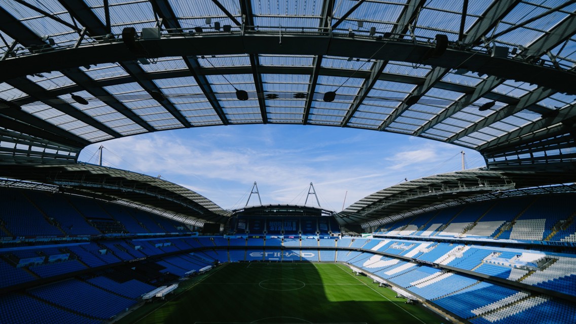 Interior view of a stadium with blue seats and a large roof structure, showing a clear sky above.