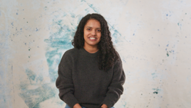 A person wearing a dark gray sweater with curly hair sitting against a creative blue and white artistic background.