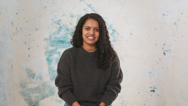 A person wearing a dark gray sweater with curly hair sitting against a creative blue and white artistic background.