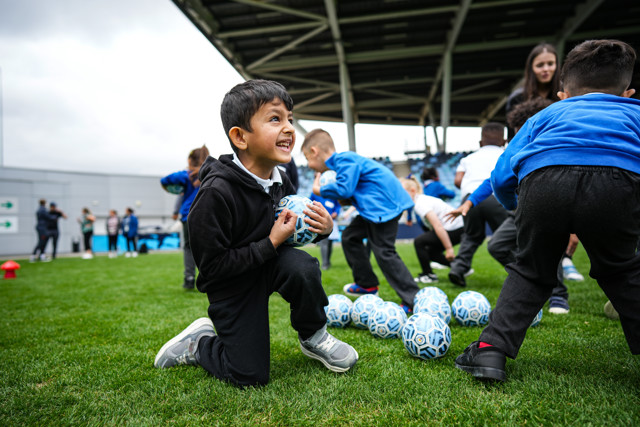 Young children engaged in soccer training, some holding blue and white soccer balls on a grassy field with a covered stadium structure in the background.