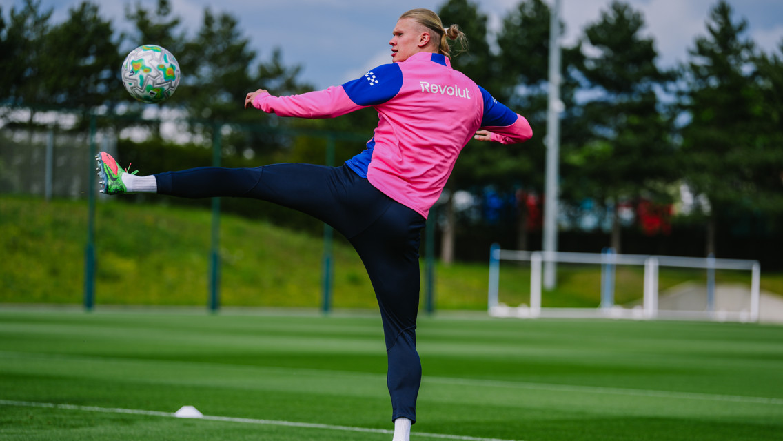 A soccer player in a pink jacket performs a high kick towards a ball during practice on a field.