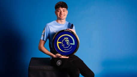 A person in a Manchester City jersey holding an ePremier League champion shield against a blue background.