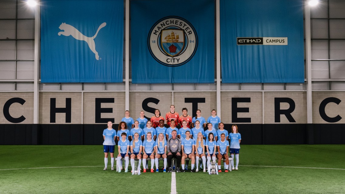 A group photo of the Manchester City women's football team in front of a Manchester City banner at the Etihad Campus indoor pitch.
