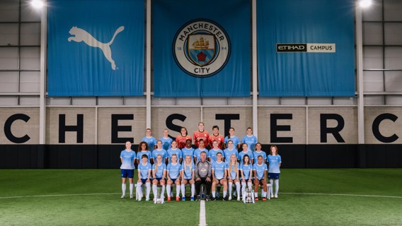 A group photo of the Manchester City women's football team in front of a Manchester City banner at the Etihad Campus indoor pitch.