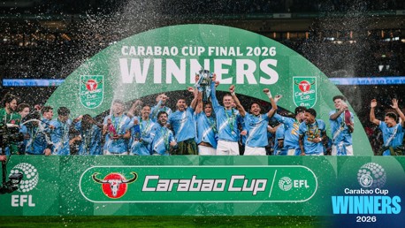 Players in light blue jerseys celebrating with a trophy under a large banner reading 'Carabao Cup Final 2026 Winners'. Confetti and celebration are visible.