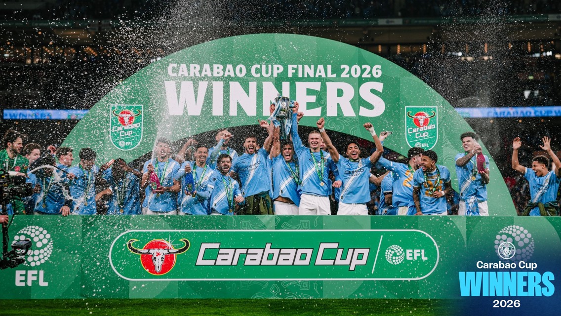 Players in light blue jerseys celebrating with a trophy under a large banner reading 'Carabao Cup Final 2026 Winners'. Confetti and celebration are visible.