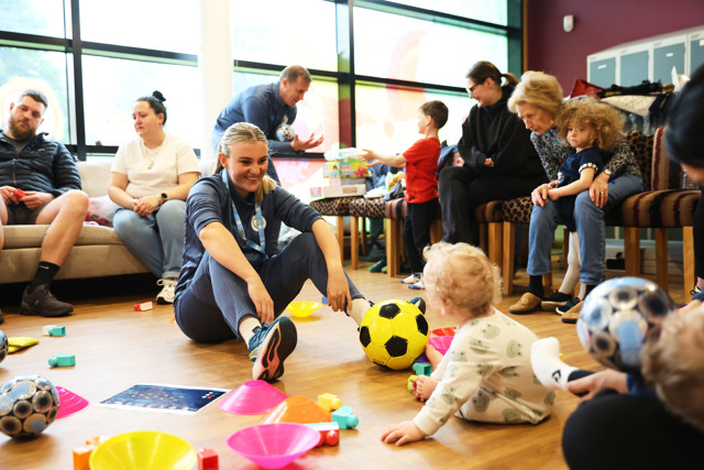 A group of adults and children are sitting and interacting in a room with toys and soccer balls scattered on the floor.