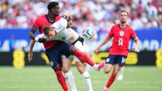 Football match scene with a fierce tackle involving players in red and white jerseys, one holding a ball.