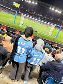 Two fans wearing Manchester City shirts with 'Maya B' and 'Ayaana' #11 in a crowded stadium, watching a football match at night.