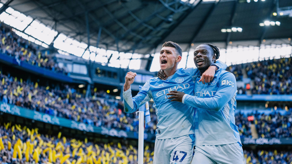 Two Manchester City players in light blue kit celebrating a goal in a stadium.