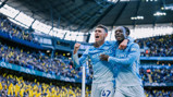 Two Manchester City players in light blue kit celebrating a goal in a stadium.
