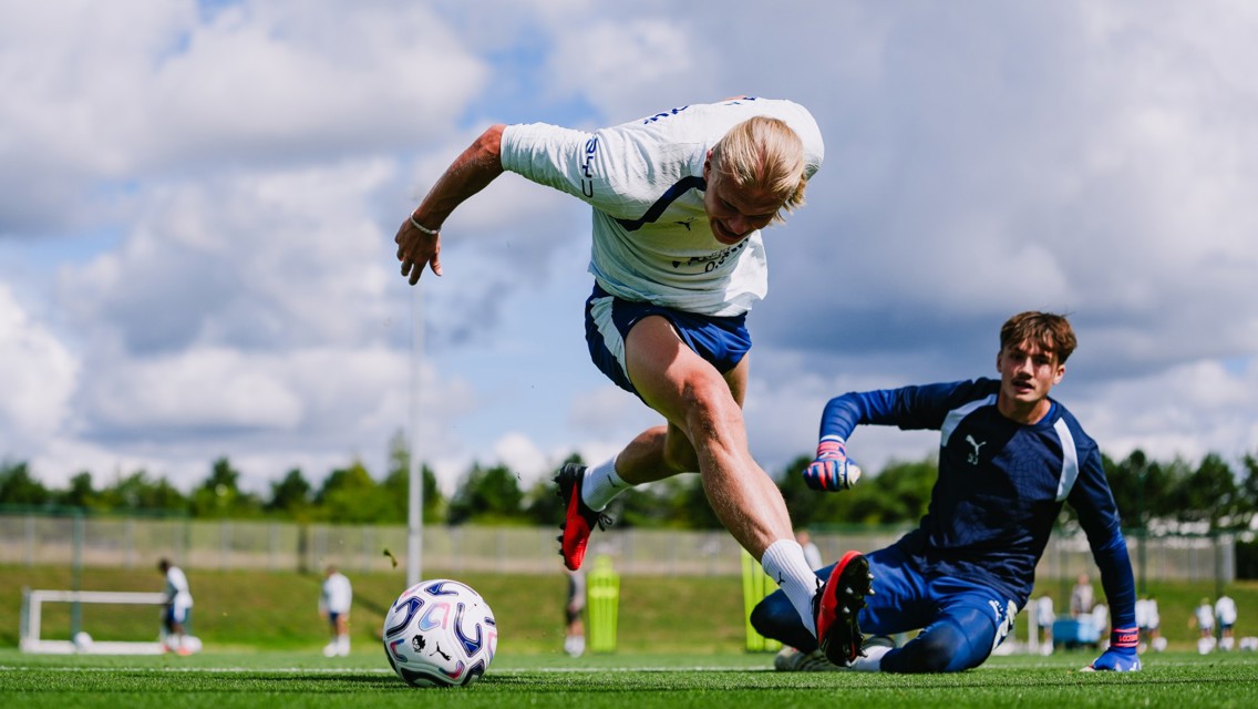 Two soccer players in action, one attempting a tackle and the other over the ball, on a green soccer field with a cloudy sky.