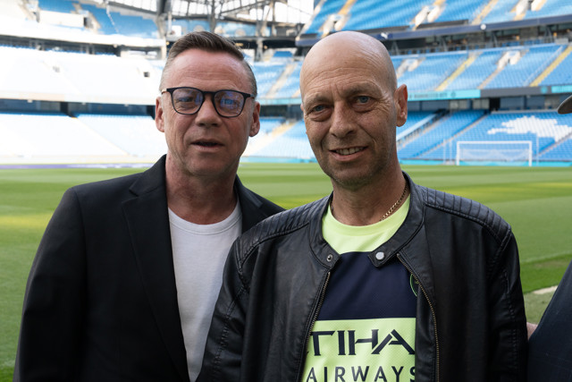 Two men standing on a football field with one wearing a Manchester City jersey inside Etihad Stadium.