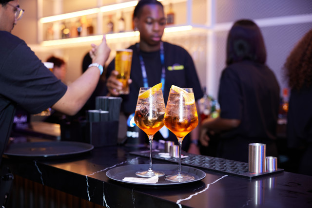 Two orange cocktails with ice and orange slices on a tray at a bar with people in the background.
