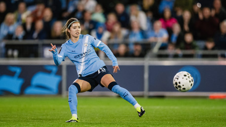 A football player wearing a Manchester City kit in action on the field, surrounded by a blurred audience.