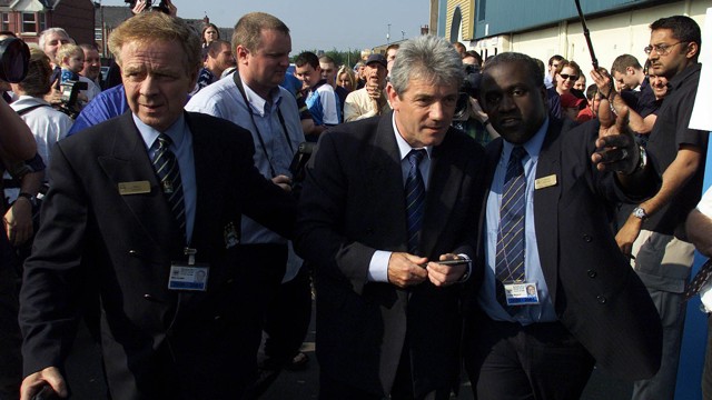 A crowd event with a security managing public figure escorted by officials, surrounded by people with cameras and passersby.