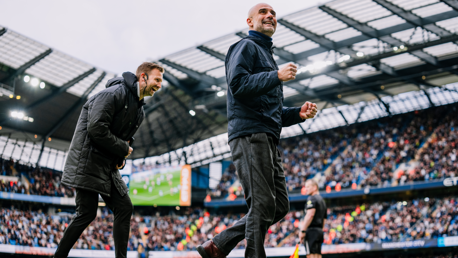 Two football coaches on the sidelines during a match at a stadium with a full crowd.