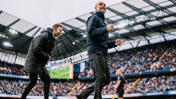Two football coaches on the sidelines during a match at a stadium with a full crowd.