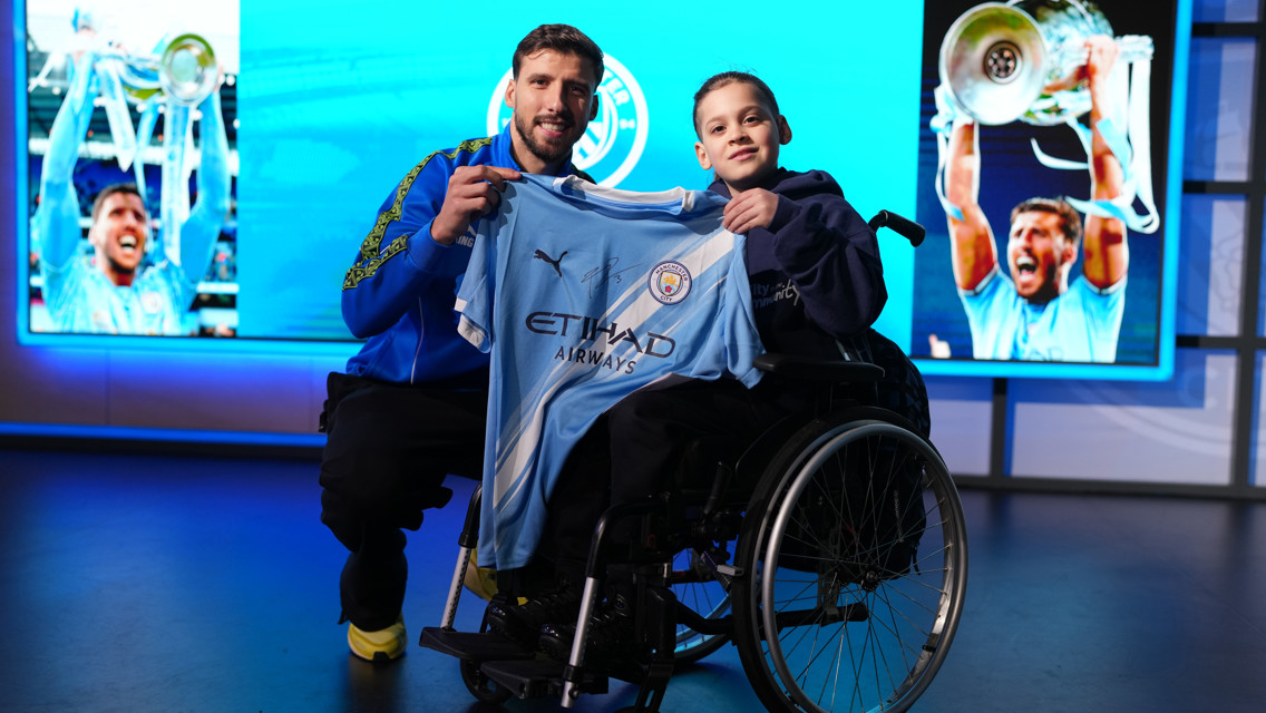 Two individuals pose with a Manchester City jersey. One person is in a wheelchair, and a large screen in the background shows team celebrations.