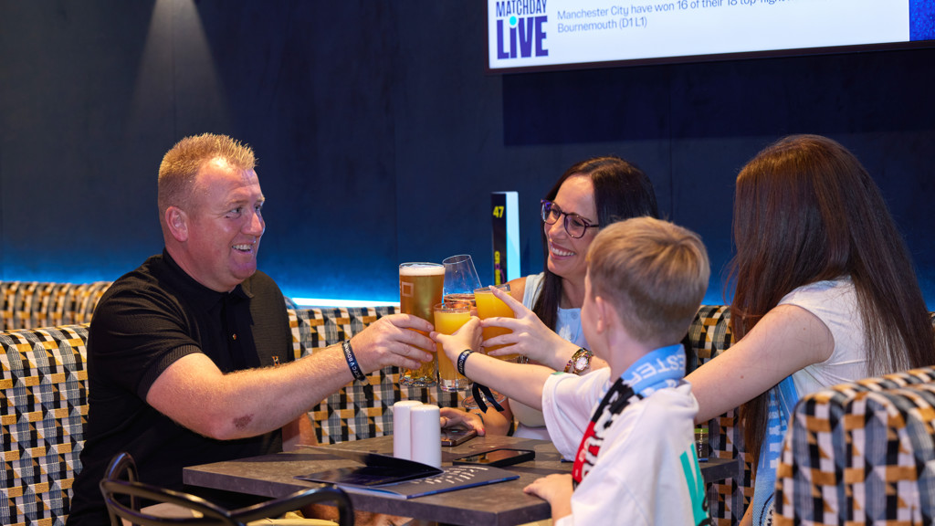 A group of people cheers with drinks in a lounge. A screen in the background shows 'Matchday Live' and data about Manchester City's matches against Bournemouth.