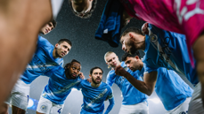 A group of Manchester City players in light blue jerseys form a huddle on a field, with the team's logo visible on their uniforms.