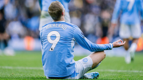 A soccer player wearing a Manchester City jersey with the number 9 and name 'Haaland' sits cross-legged on the field in a meditative posture during a match.