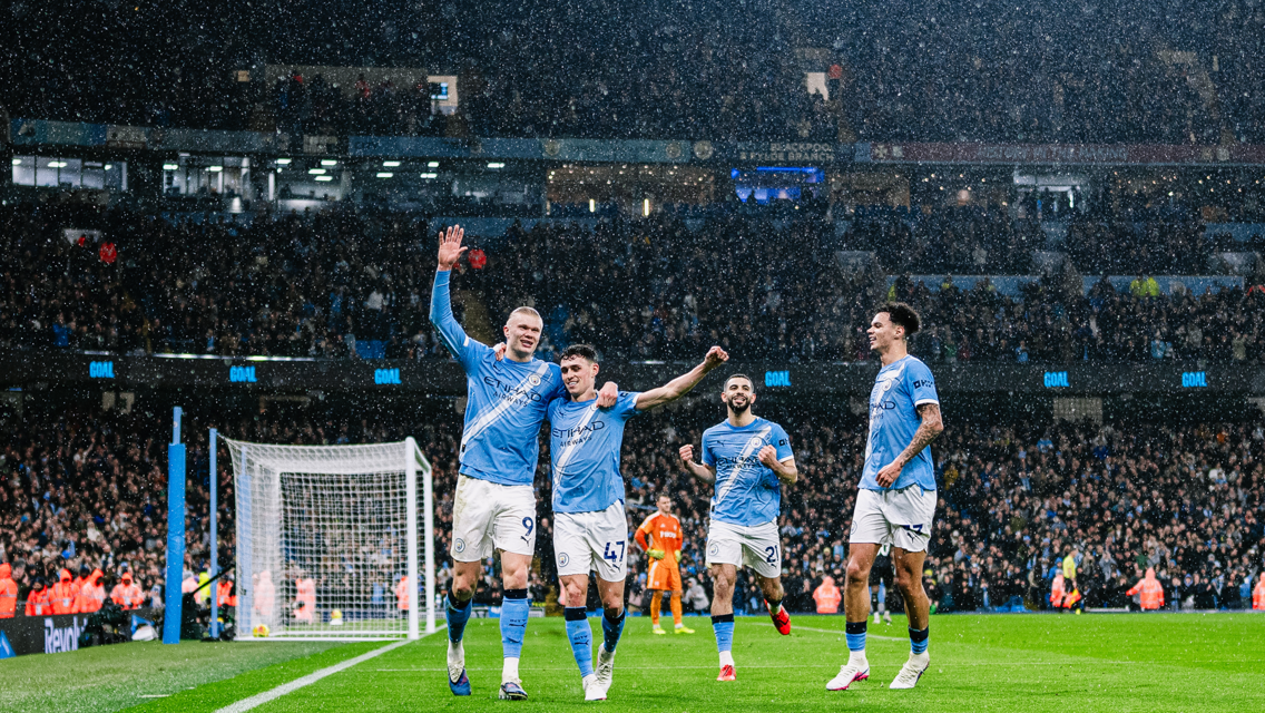 Football players wearing light blue kits celebrating a goal in a stadium under the rain, with a crowd in the background.