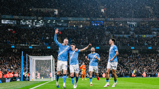 Football players wearing light blue kits celebrating a goal in a stadium under the rain, with a crowd in the background.