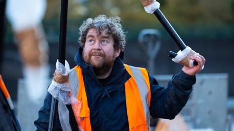 A construction worker with curly hair and a beard wearing a high-visibility orange jacket and holding poles wrapped in bubble wrap outdoors.