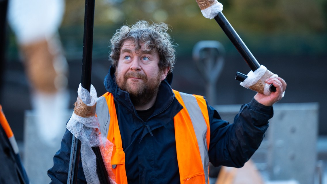 A construction worker with curly hair and a beard wearing a high-visibility orange jacket and holding poles wrapped in bubble wrap outdoors.