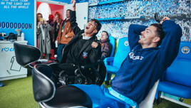 Group of people inside a room with Manchester City branding. Two adults sit in blue chairs, surrounded by memorabilia and club decorations. Others stand observing the interaction.