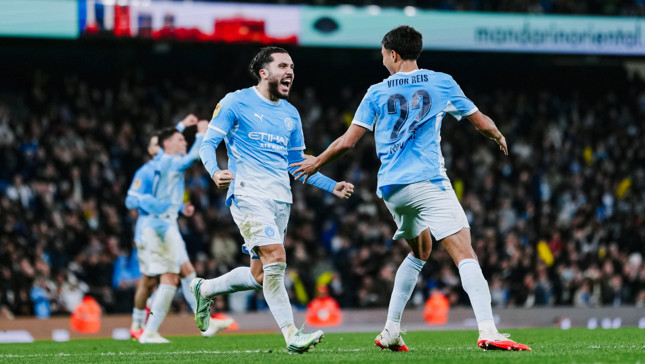 Two Manchester City players wearing blue uniforms celebrate on the field, with a crowd in the background.