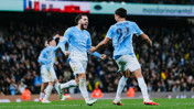 Two Manchester City players wearing blue uniforms celebrate on the field, with a crowd in the background.