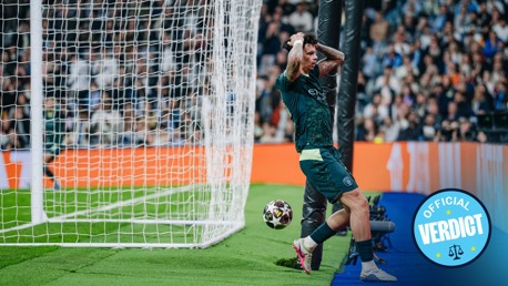 Football player in a dark uniform reacts with hands on head near the goal post after an unsuccessful attempt to score, with the ball rolling past and fans in the background.