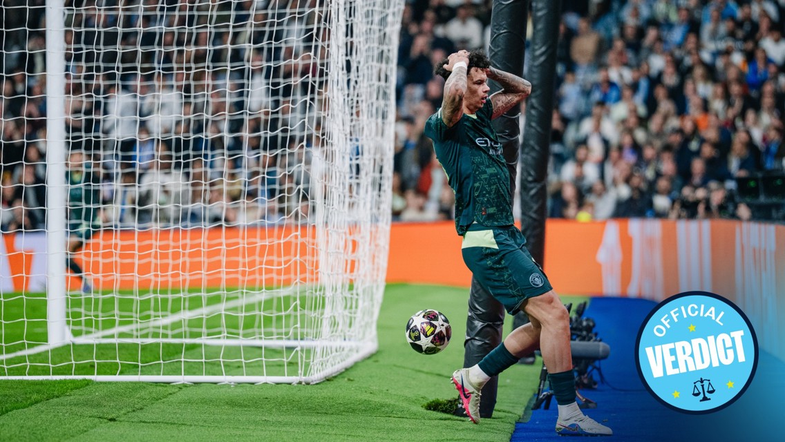 Football player in a dark uniform reacts with hands on head near the goal post after an unsuccessful attempt to score, with the ball rolling past and fans in the background.