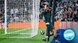 Football player in a dark uniform reacts with hands on head near the goal post after an unsuccessful attempt to score, with the ball rolling past and fans in the background.