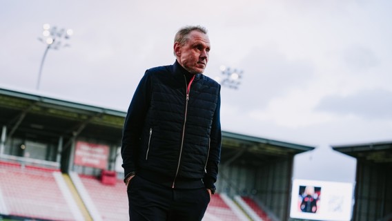 A man wearing a black jacket standing in a stadium with empty seats in the background.