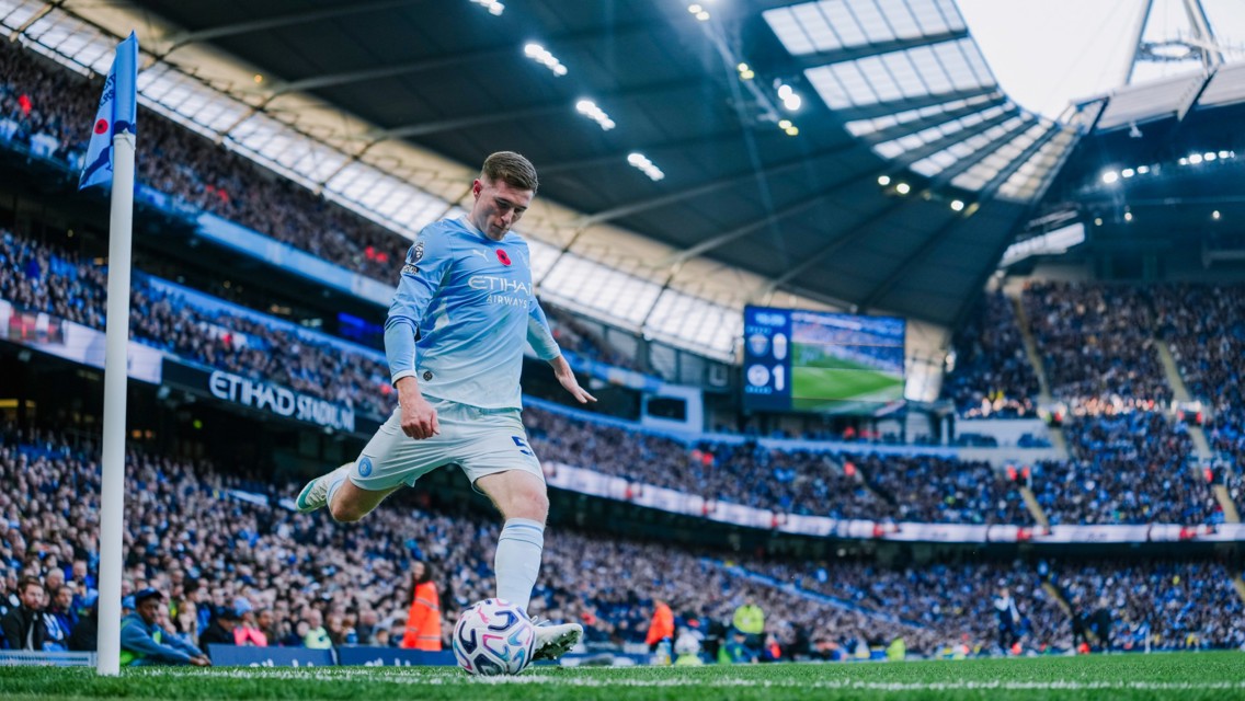A Manchester City player taking a corner kick at Etihad Stadium during a match. The player is wearing a light blue kit and the stadium is filled with a large crowd of spectators.